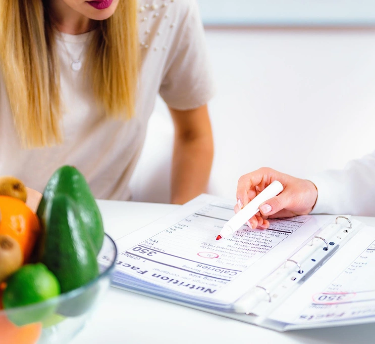A nutritionist points to a food chart in a binder as a person listens, conveying a sense of guidance. A bowl of fresh fruits and vegetables is nearby.