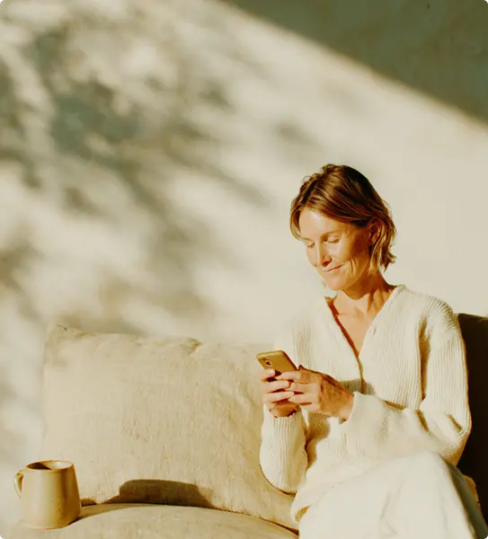 Woman sitting on a couch in warm sunlight, using a smartphone.
