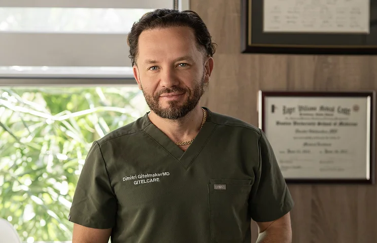 Dr. Dimitri Gitelmaker MD in olive scrubs standing in his medical office with diplomas on the wall
