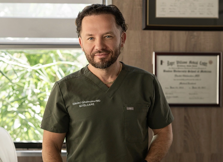 Portrait of Dr. Dimitri Gitelmaker in olive green GitelCare scrubs standing in his office beside framed medical diplomas